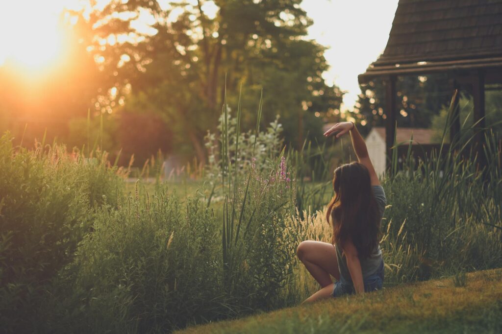 A woman sitting in a garden, stretching her arm under the warm sunset light, embracing nature's tranquility.